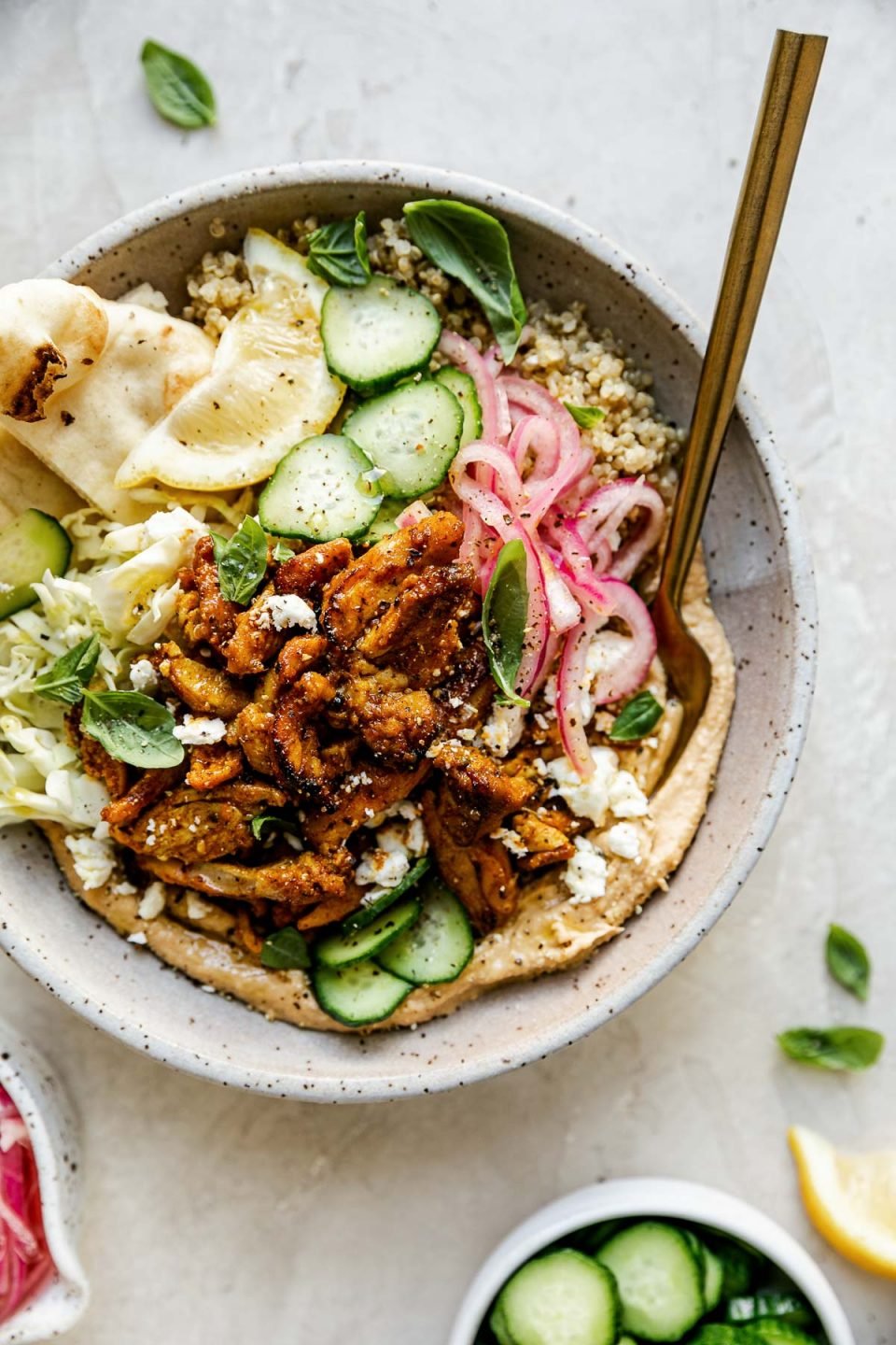 Chicken Shawarma Hummus Bowl with quinoa, cucumbers, pickled red onion, shredded cabbage, & naan bread shown in a speckled gray ceramic bowl. The bowl sits atop a creamy cement surface, surrounded by lemon wedges, fresh basil leaves, a small bowl of pickled red onions, & a small bowl of sliced cucumbers.