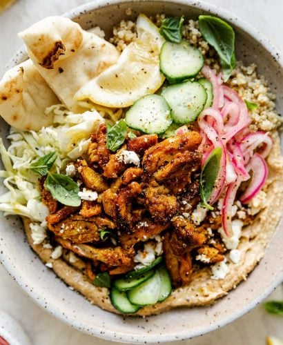 Chicken Shawarma Hummus Bowl with quinoa, cucumbers, pickled red onion, shredded cabbage, & naan bread shown in a speckled gray ceramic bowl. The bowl sits atop a creamy cement surface, surrounded by lemon wedges, fresh basil leaves & a small bowl of pickled red onions.