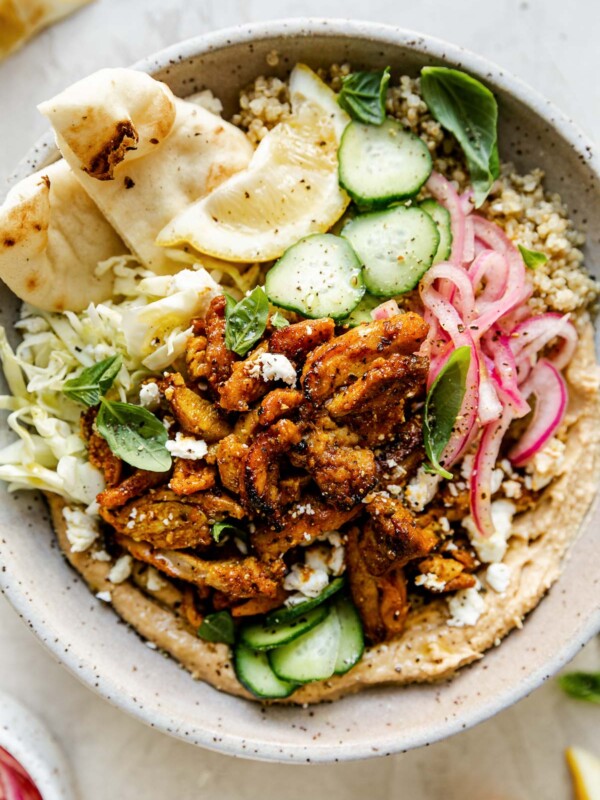 Chicken Shawarma Hummus Bowl with quinoa, cucumbers, pickled red onion, shredded cabbage, & naan bread shown in a speckled gray ceramic bowl. The bowl sits atop a creamy cement surface, surrounded by lemon wedges, fresh basil leaves & a small bowl of pickled red onions.