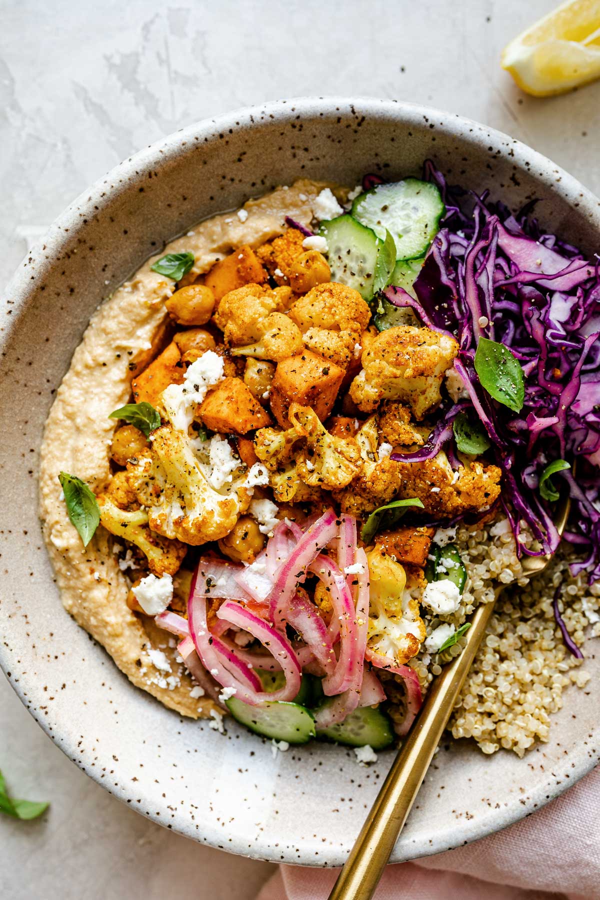 Vegan shawarma hummus bowl, with roasted cauliflower & sweet potato shawarma, thinly sliced cucumber, pickled red onions, red cabbage, & quinoa. The bowl sits atop a creamy cement surface next to lemon wedges & a pink linen napkin.