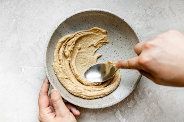 A woman's hands spreading hummus on the bottom of a speckled gray ceramic bowl with a large spoon. The bowl sits atop a creamy cement surface.