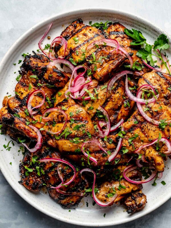 Grilled Shawarma marinated chicken thighs shown on a white speckled plate atop a light blue surface. The chicken is garnished with fresh herbs & ribbons of pickled red onion.