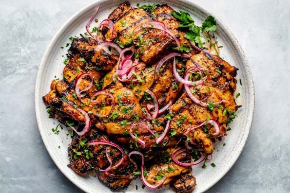 Grilled Shawarma marinated chicken thighs shown on a white speckled plate atop a light blue surface. The chicken is garnished with fresh herbs & ribbons of pickled red onion.