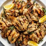 Grilled Lemon & Herb marinated chicken thighs shown on a white speckled plate atop a light blue surface. The chicken is garnished with fresh herbs & lemon wedges.