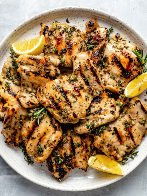 Grilled Lemon & Herb marinated chicken thighs shown on a white speckled plate atop a light blue surface. The chicken is garnished with fresh herbs & lemon wedges.