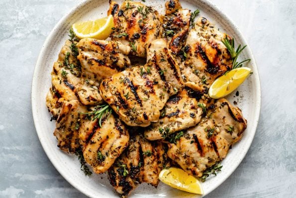 Grilled Lemon & Herb marinated chicken thighs shown on a white speckled plate atop a light blue surface. The chicken is garnished with fresh herbs & lemon wedges.