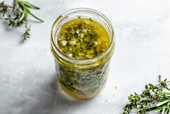 Lemon herb marinade shown in a small ball jar, sitting atop a light blue surface with sprigs of fresh rosemary & thyme in the foreground & in the background.