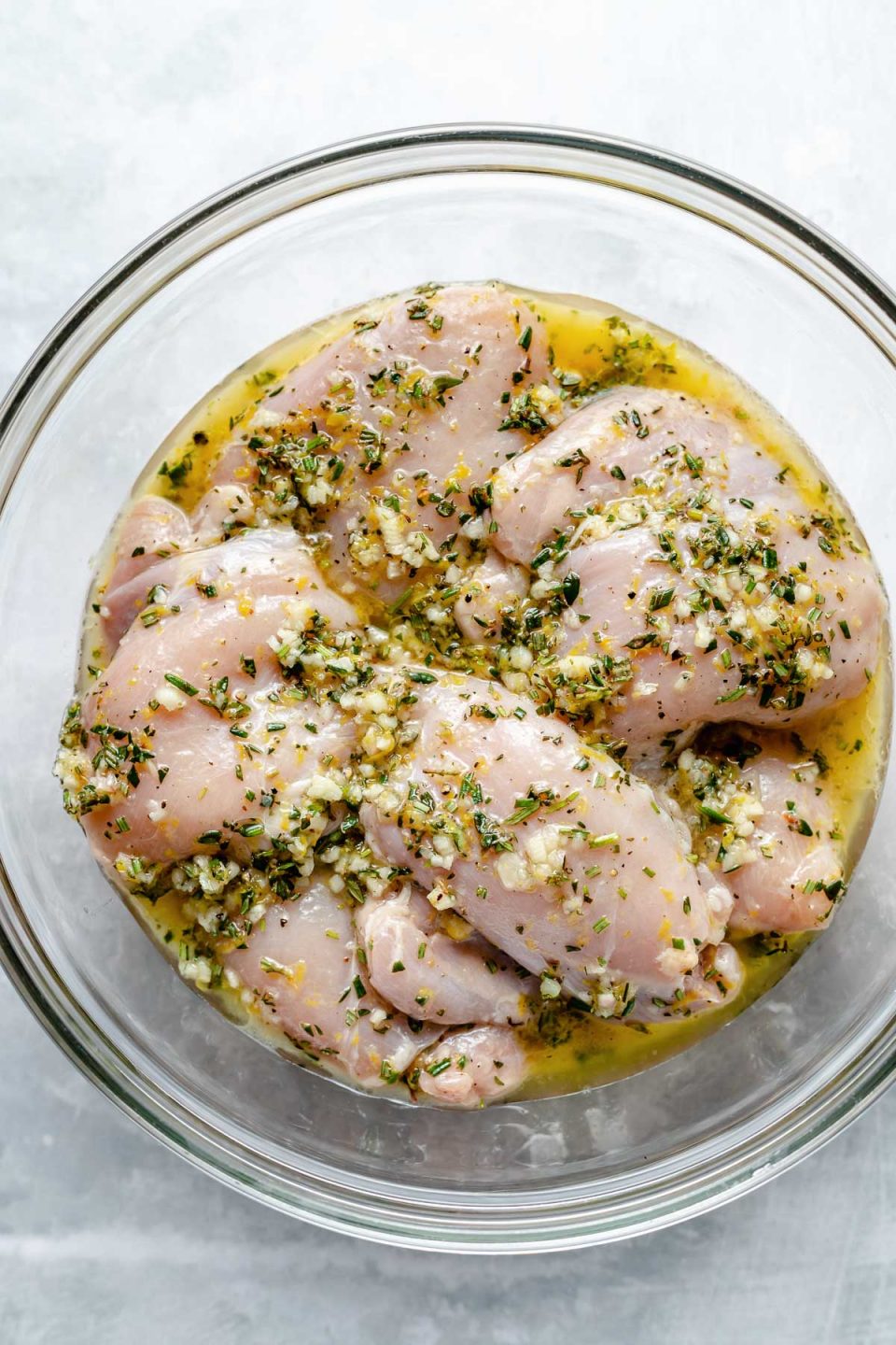 Chicken thighs in a large glass mixing bowl, marinating in Lemon Herb marinade. The bowl sits atop a light blue surface.