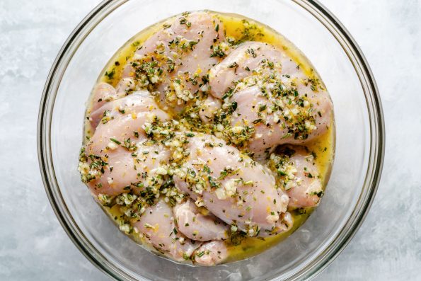Chicken thighs in a large glass mixing bowl, marinating in Lemon Herb marinade. The bowl sits atop a light blue surface.