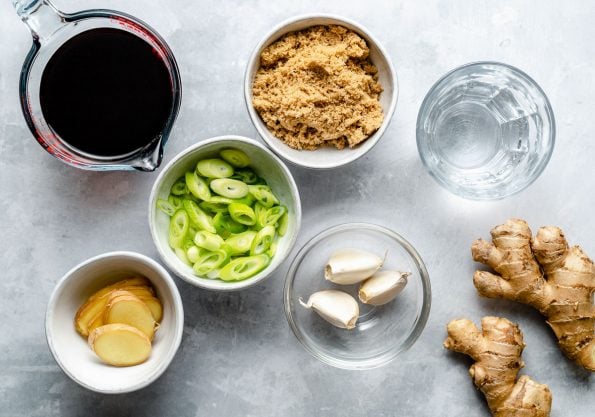 Teriyaki marinade ingredients arranged on a light blue surface: soy sauce, water, brown sugar, garlic, green onions, & fresh ginger.