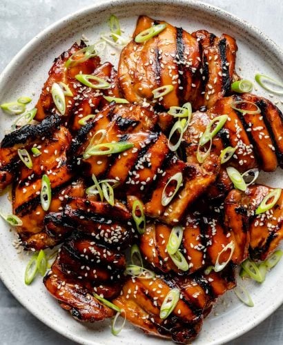 Grilled Teriyaki chicken thighs shown on a white speckled plate atop a light blue surface. The chicken is garnished with sliced green onion & sesame seeds.