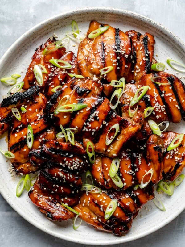 Grilled Teriyaki chicken thighs shown on a white speckled plate atop a light blue surface. The chicken is garnished with sliced green onion & sesame seeds.