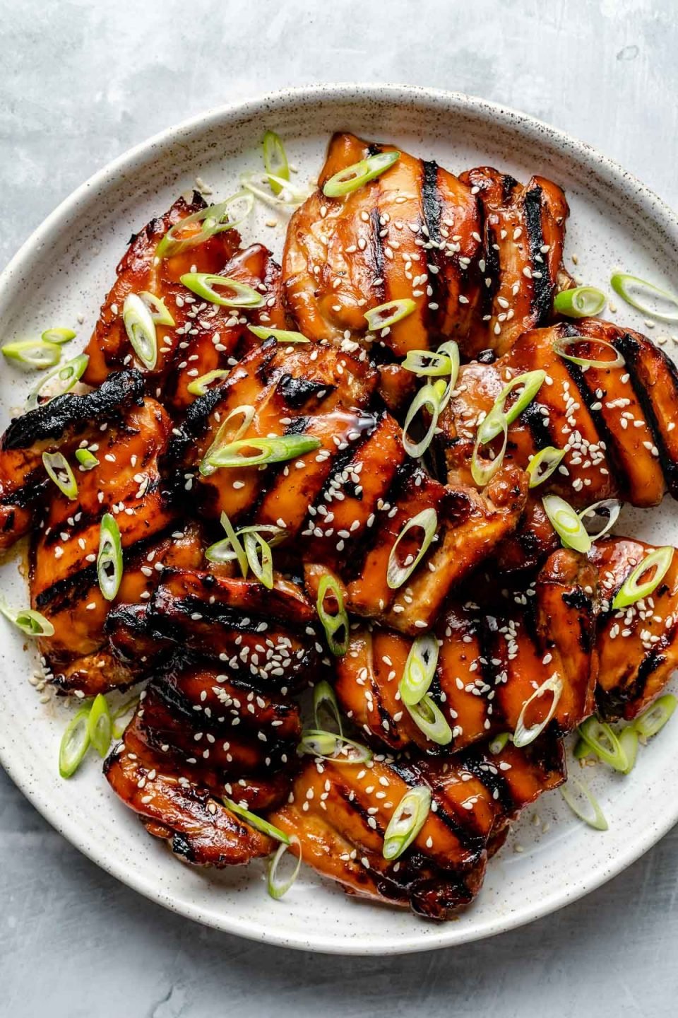 Grilled Teriyaki chicken thighs shown on a white speckled plate atop a light blue surface. The chicken is garnished with sliced green onion & sesame seeds.