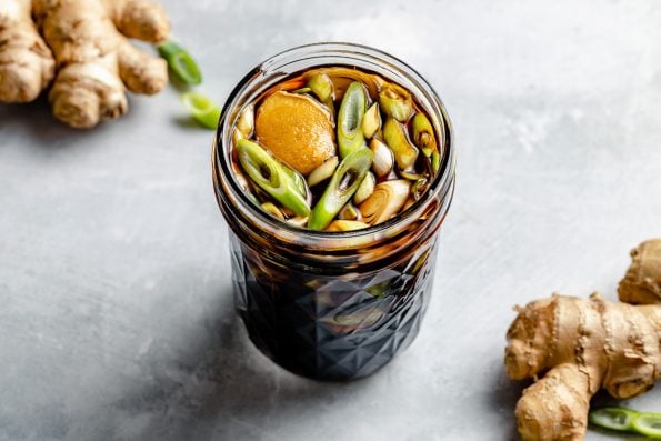Teriyaki marinade shown in a small ball jar, sitting atop a light blue surface with green onion & fresh ginger in the foreground & the background.