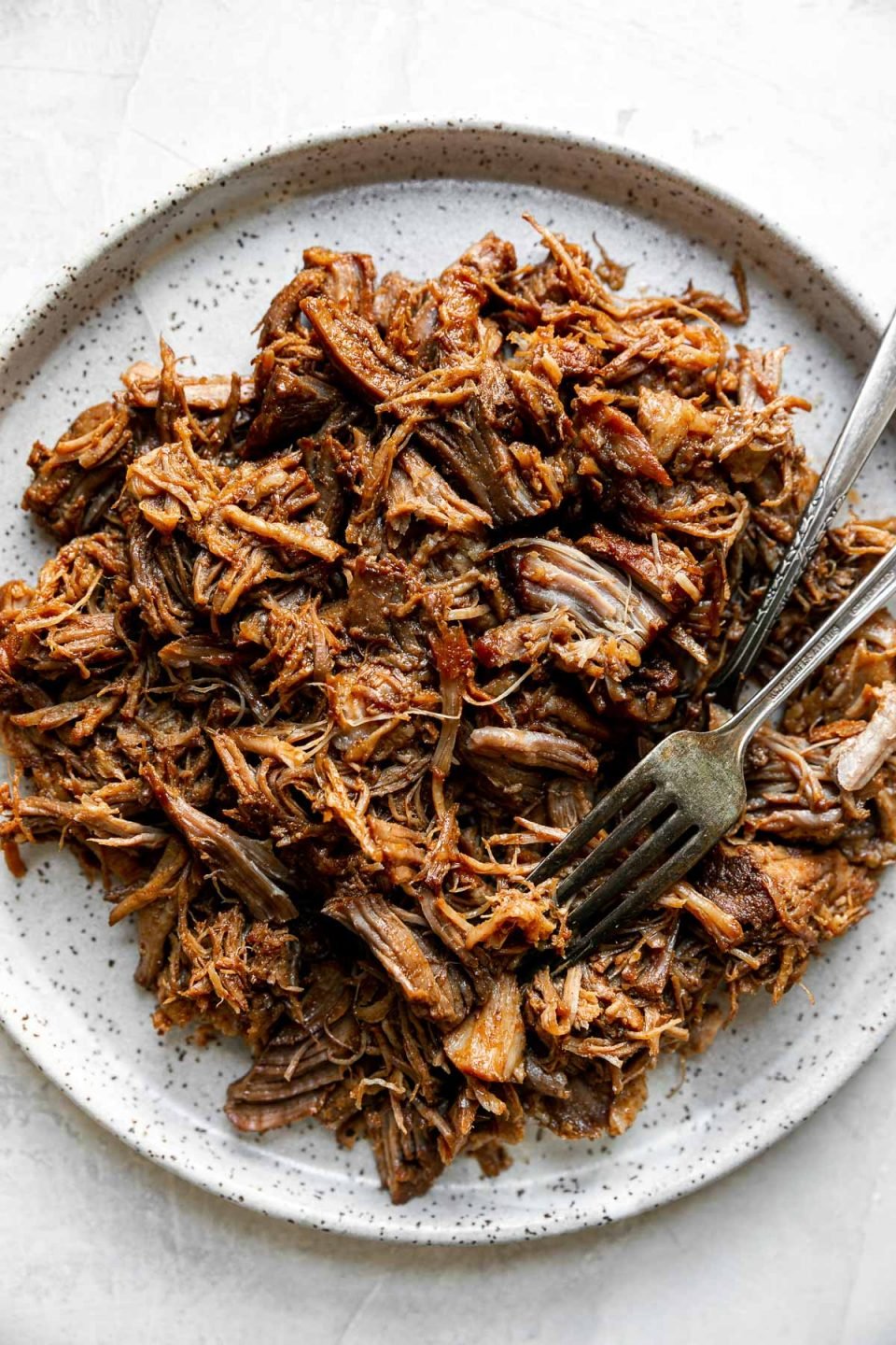 Close up of bbq pulled pork shown on a speckled ceramic plate, with 2 forks. The plate sits atop a creamy cement surface.