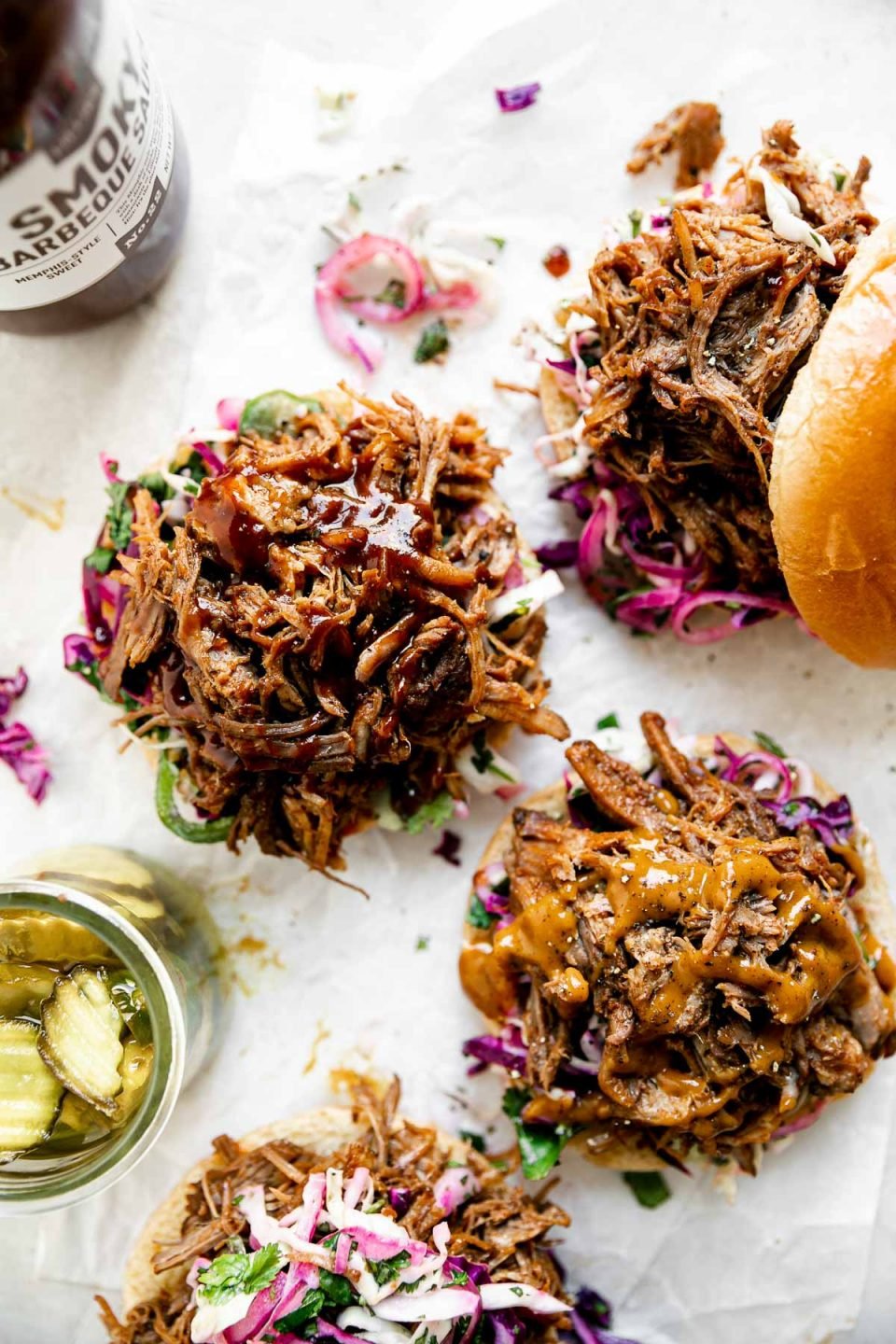 Easy pulled pork shown on 4 open face sandwiches. The pork sits atop buns & slaw on a white surface, alongside a jar of pickles & BBQ sauce.