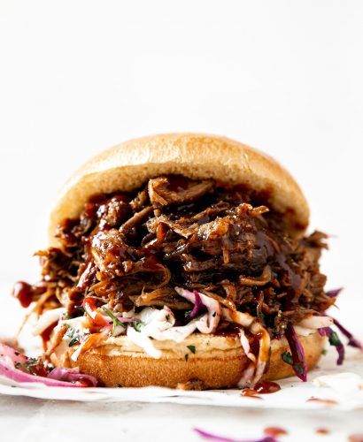 Straight-on view of pulled pork sandwich. Pulled pork & slaw on a brioche bun, atop a creamy white surface.