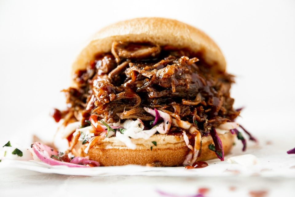 Straight-on view of pulled pork sandwich. Pulled pork & slaw on a brioche bun, atop a creamy white surface.