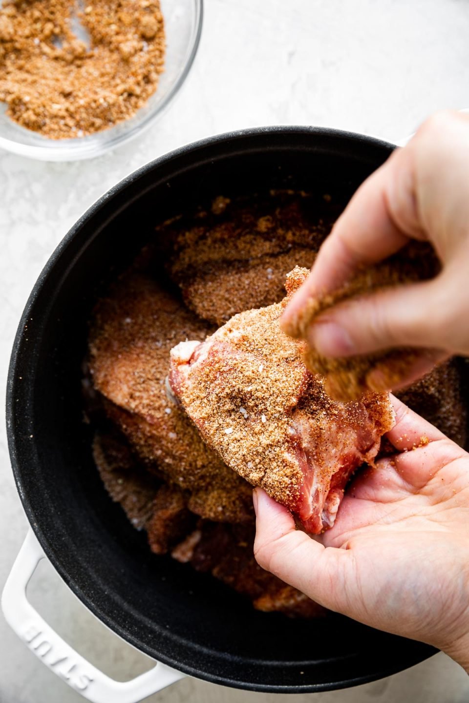 A woman's hands shown sprinkling bbq dry rub on pork shoulder over a white Staub dutch oven. The dutch oven sits atop a creamy cement surface, next to a bowl of BBQ dry rub.