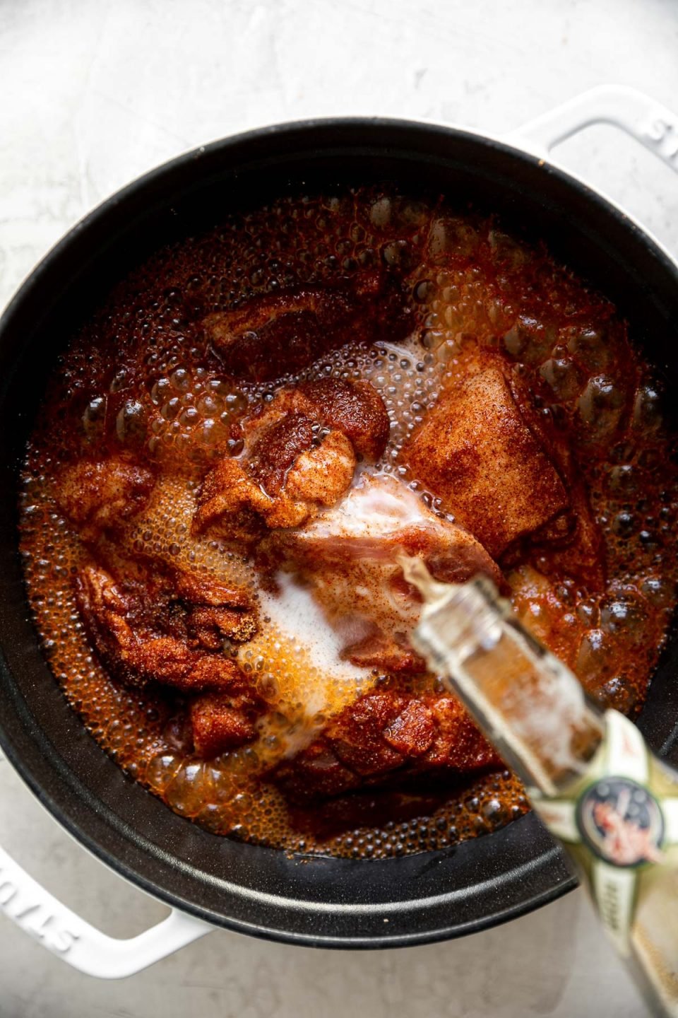 Seasoned pork shown in large white dutch oven atop a creamy cement surface. A bottle of Miller High Life is shown pouring into the dutch oven, over the pork.