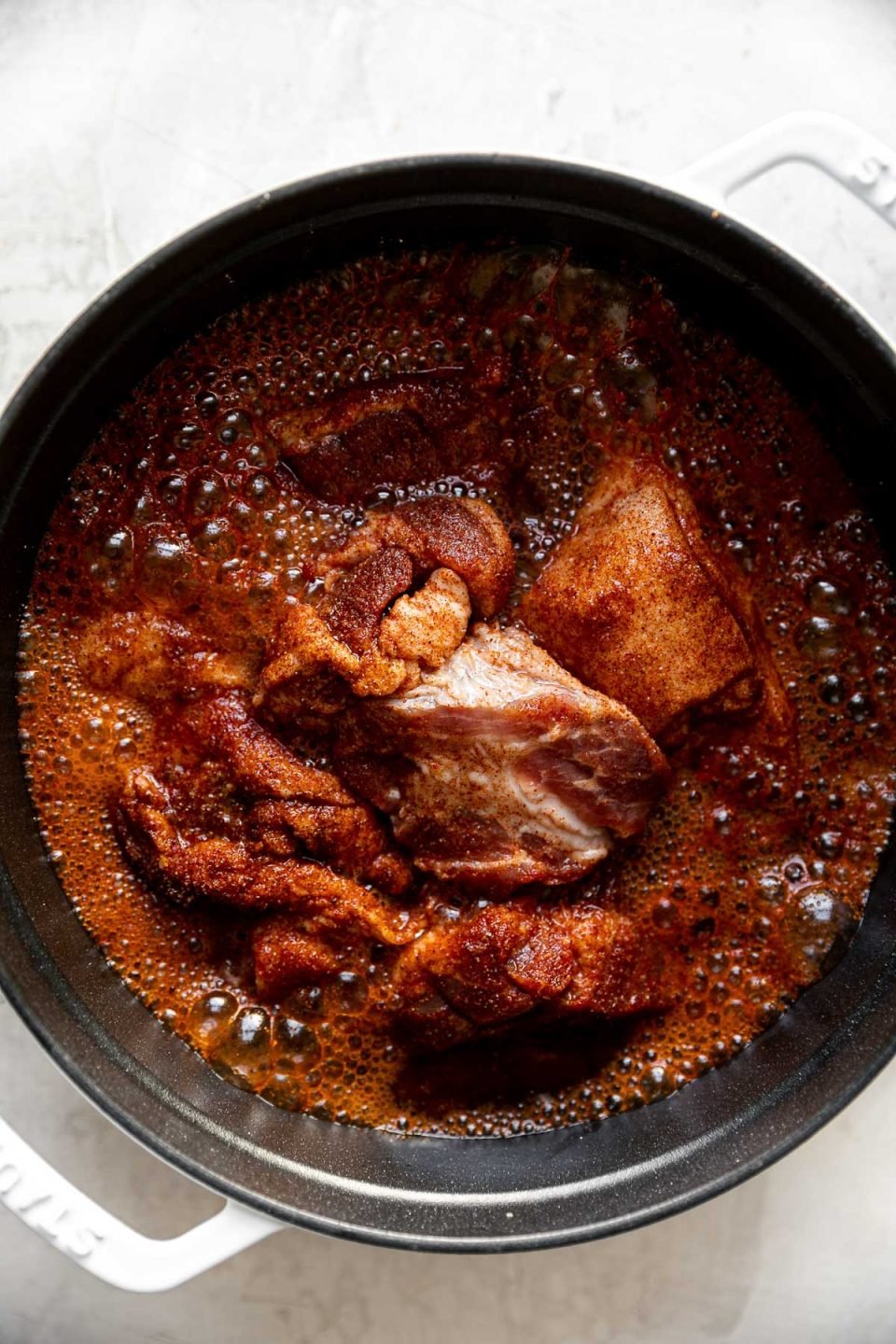 Seasoned pork shown in large white dutch oven atop a creamy cement surface.