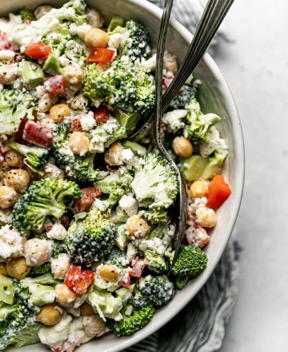 Healthy broccoli salad in a large white serving bowl atop a gray & white striped linen napkin on a light gray surface. 2 rustic serving spoons are nestled into the broccoli salad.
