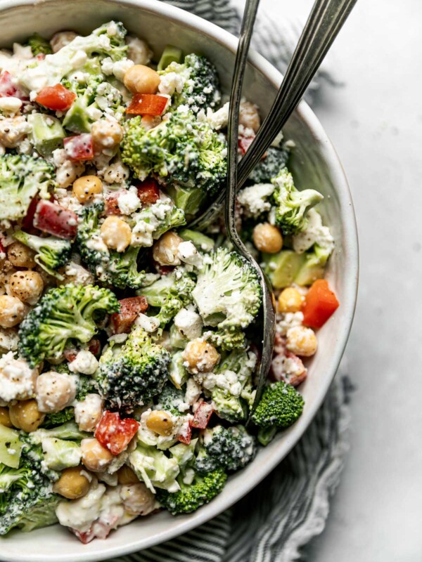 An overhead shot of broccoli salad in a large white serving bowl atop a gray and white striped linen napkin on a light gray surface. 2 rustic serving spoons are in the serving bowl.