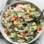 Healthy broccoli salad in a large white serving bowl atop a gray & white striped linen napkin on a light gray surface. 2 rustic serving spoons are in the serving bowl.