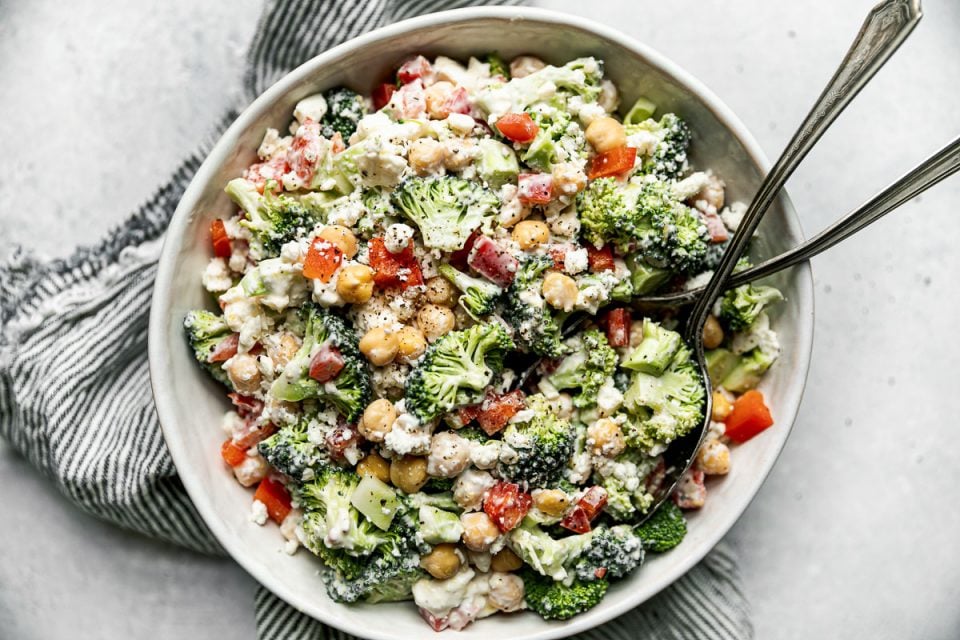 Healthy broccoli salad in a large white serving bowl atop a gray & white striped linen napkin on a light gray surface. 2 rustic serving spoons are in the serving bowl.