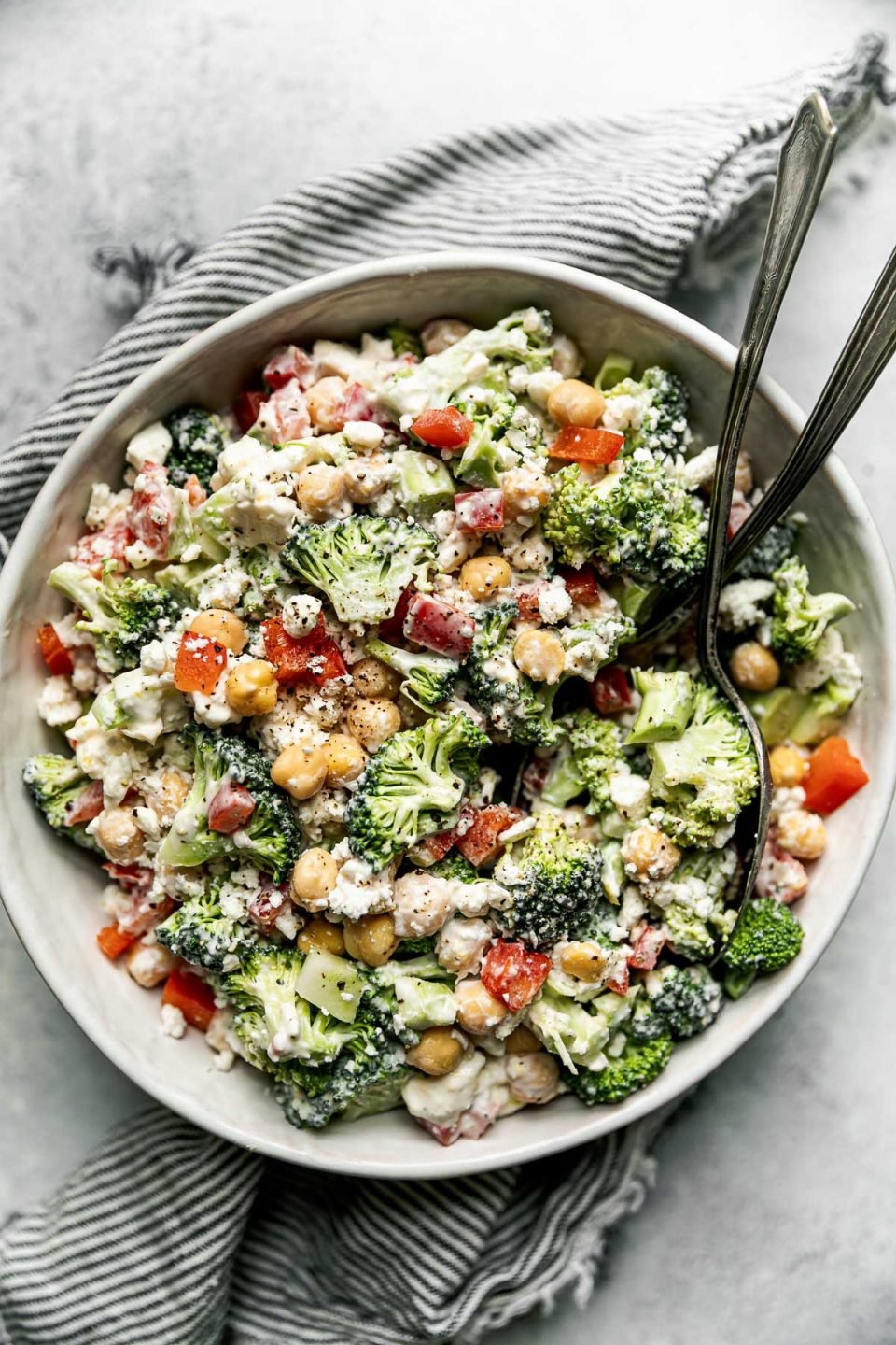 Healthy broccoli salad in a large white serving bowl atop a gray & white striped linen napkin on a light gray surface. 2 rustic serving spoons are nestled into the broccoli salad.
