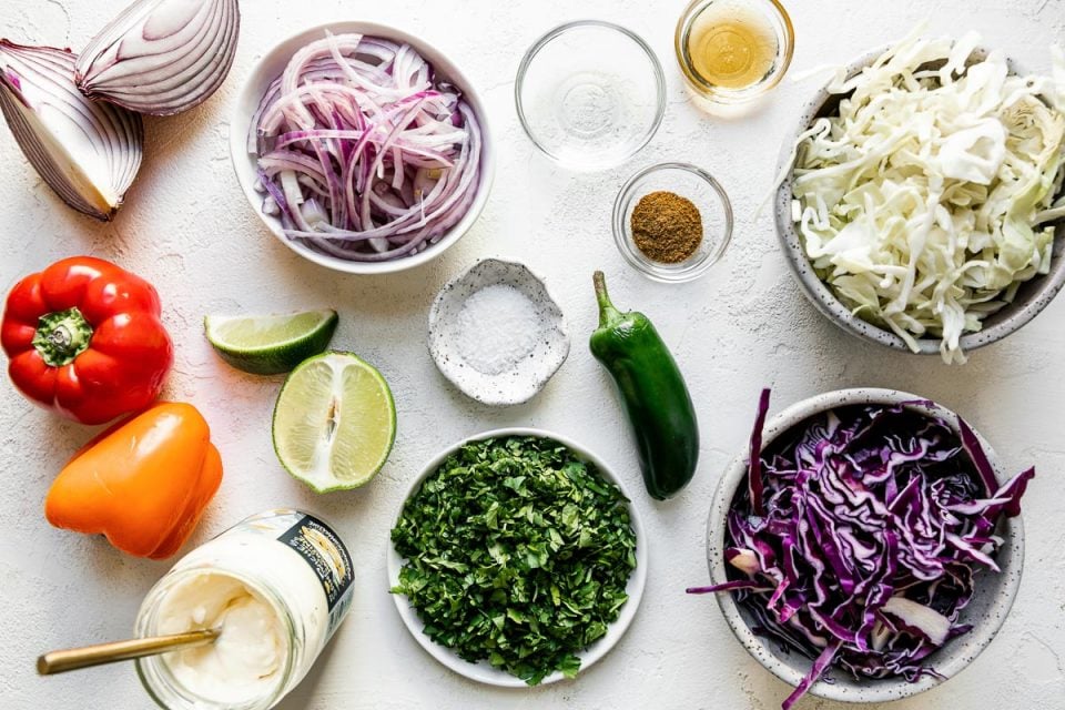 Easy slaw ingredients arranged on a textured white surface – red onion, bell pepper, lime, mayonnaise, chopped cilantro, salt, cumin, vinegar, agave, shredded cabbage & jalapeno pepper.