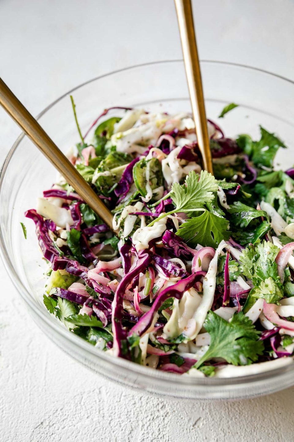 Side angle of easy slaw shown in a large glass mixing bowl atop a textured white surface with 2 gold forks inserted in it.