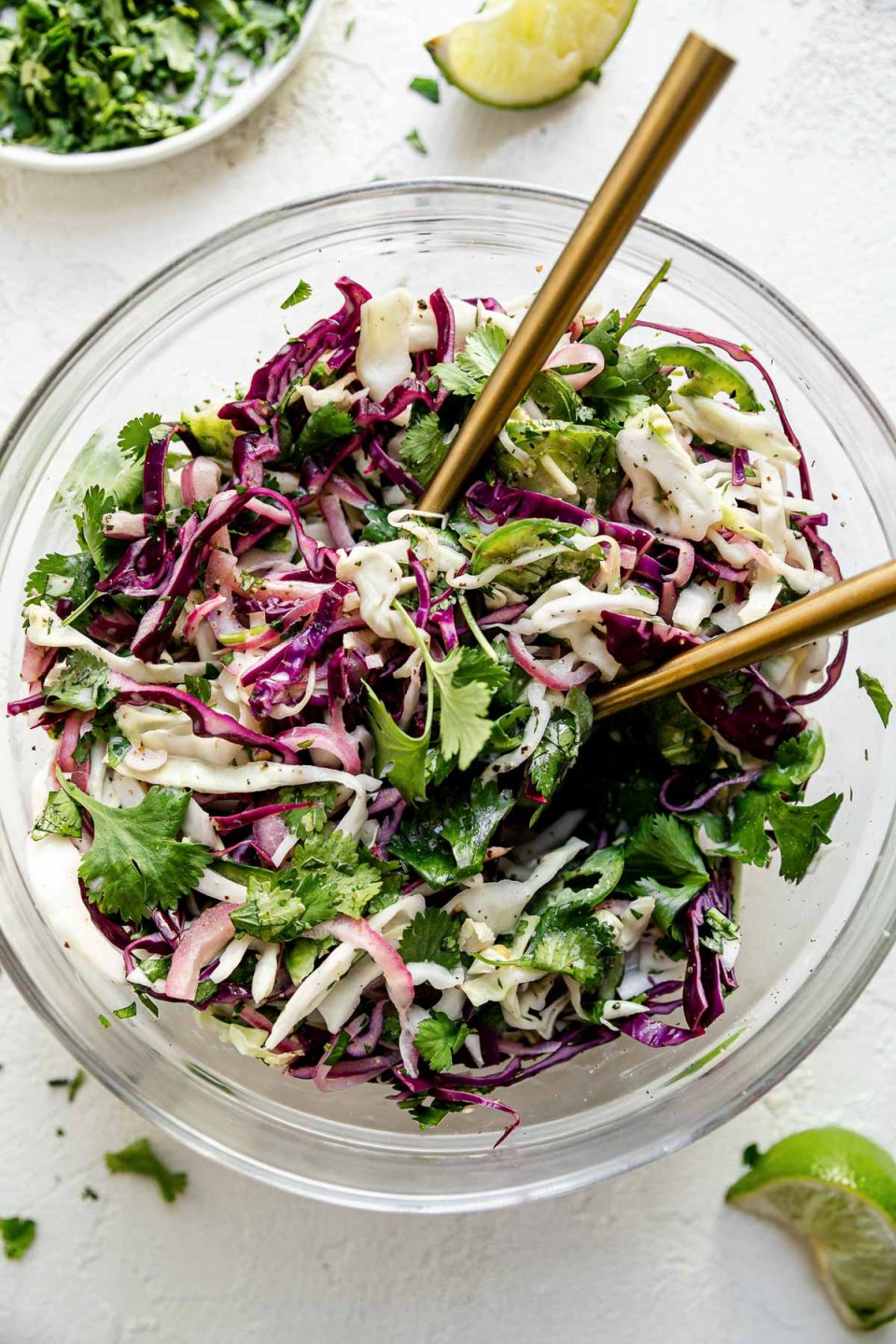 Easy slaw shown in a large glass mixing bowl atop a textured white surface with 2 gold forks inserted in it. Surrounding the bowl are chopped cilantro leaves, a small plate of chopped cilantro, & a spent lime wedge.