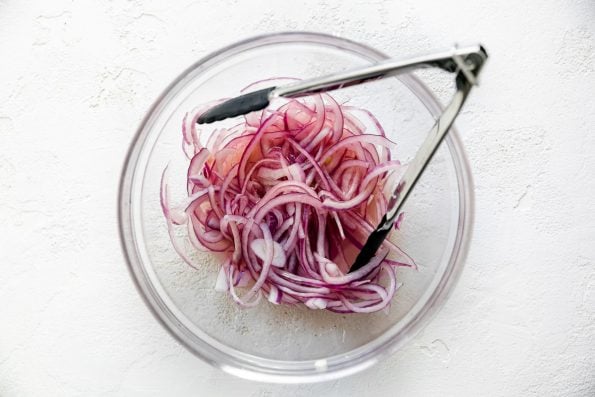 How to make simple slaw, Step 1: Thinly sliced red onion in a clear glass bowl soaking in lime juice. There is a small pair of tongs in the bowl with the onions, which sits atop a textured white surface.