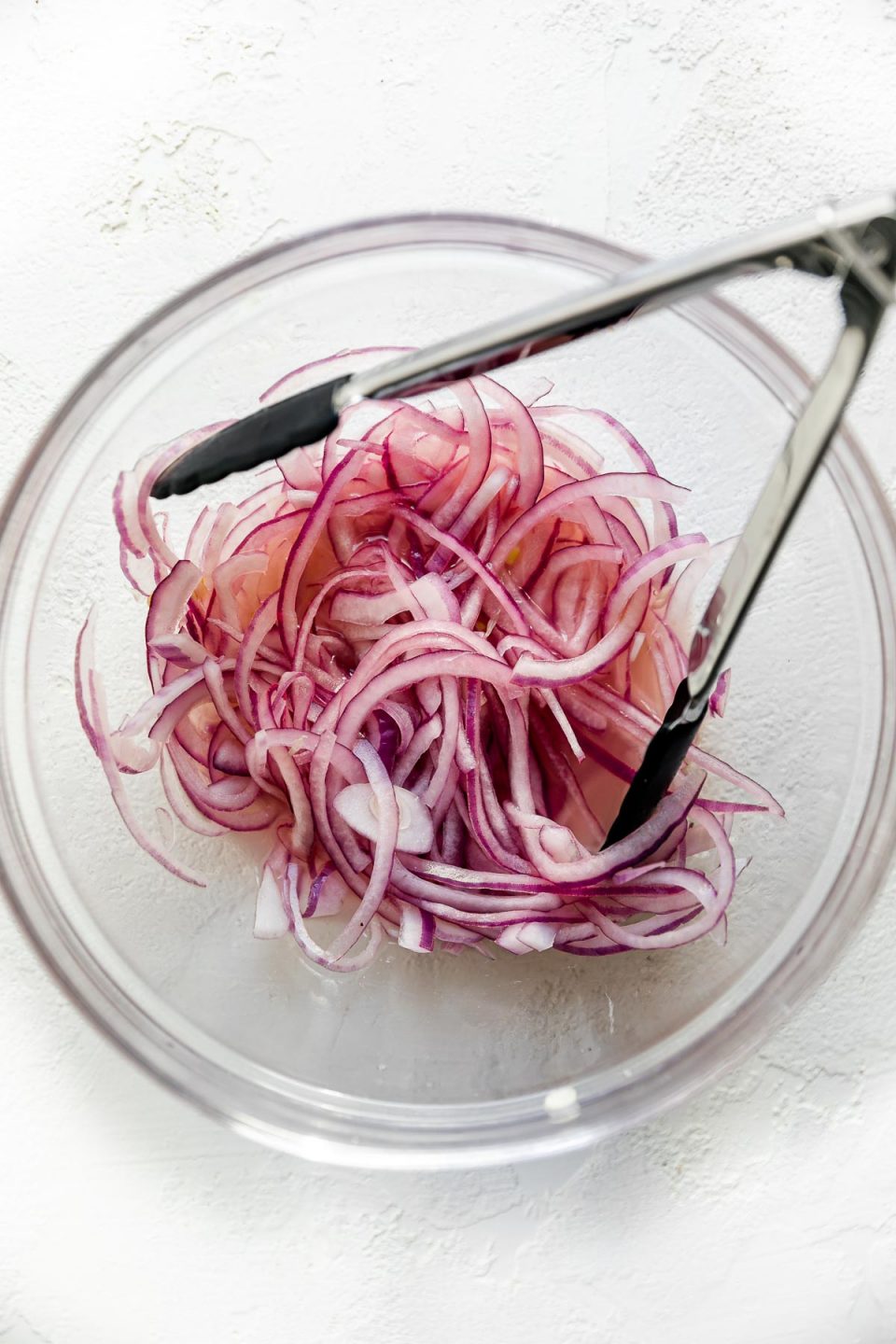 Thinly sliced red onion in a clear glass bowl soaking in lime juice. There is a small pair of tongs in the bowl with the onions, which sits atop a textured white surface.