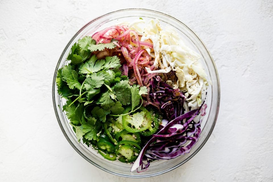 How to make simple slaw, Step 3: Simple slaw ingredients in a large glass mixing bowl – cabbage, lime-soaked onions, cilantro leaves, & jalapeno peppers. The bowl sits atop a textured white surface.