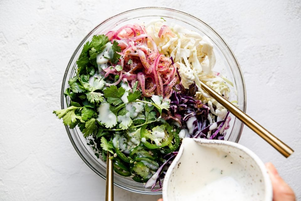 How to make simple slaw, Step 3: Pouring slaw dressing into large mixing bowl with slaw ingredients. There are 2 gold forks inserted in the bowl, which sits atop a white textured surface.