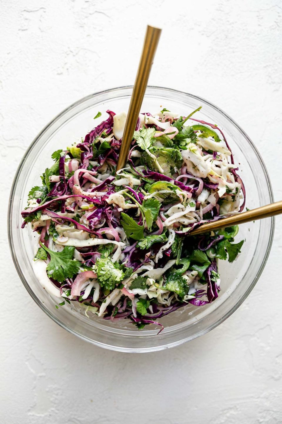 Easy slaw shown in a large glass mixing bowl atop a textured white surface with 2 gold forks inserted in it.