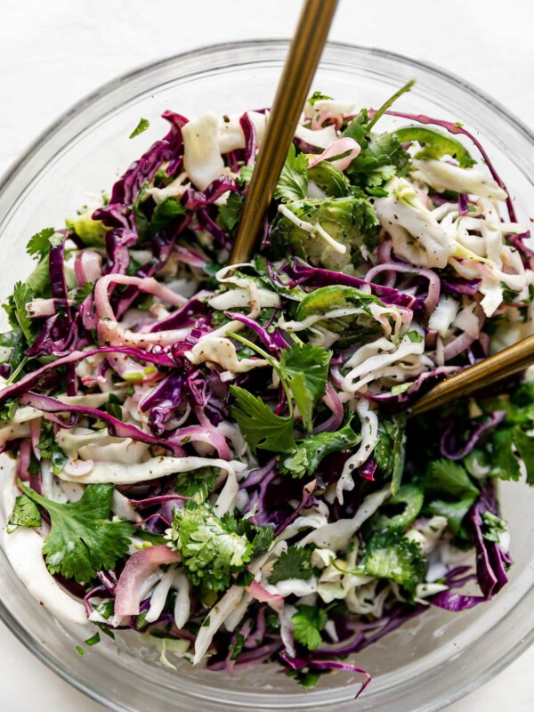 Close up of easy slaw shown in a large glass mixing bowl atop a textured white surface with 2 gold forks inserted in it.