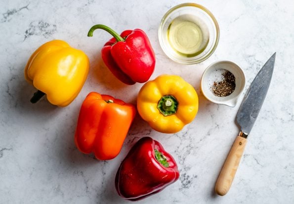 Grilled bell pepper ingredients arranged on a white & grey marble surface - yellow, orange, & red bell peppers, avocado oil, kosher salt, & ground black pepper. A pairing knife rests on the surface next to the ingredients.