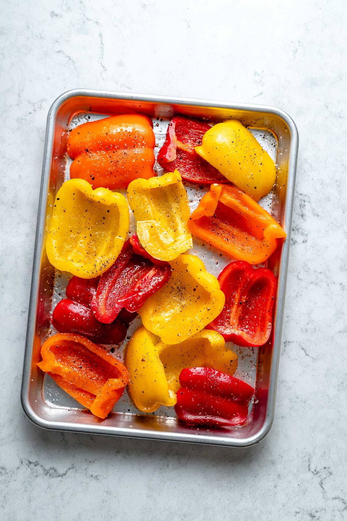 Multiple slices of fresh & raw yellow, orange, & red bell peppers seasoned with avocado oil, kosher salt, & ground black pepper arranged on an aluminum baking sheet. The baking sheet sits on top of a white & gray marble surface.
