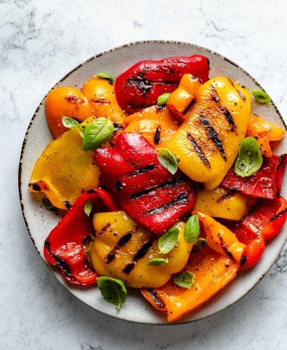 Slices of grilled bell pepper with char marks arranged on a ceramic plate. The grilled bell peppers are garnished with ground black pepper, kosher salt, & chopped fresh herbs. The plate sits on top of a white & gray marble surface.