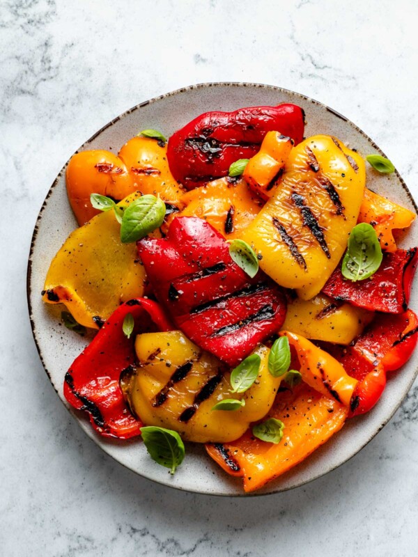 Slices of grilled bell pepper with char marks arranged on a ceramic plate. The grilled bell peppers are garnished with ground black pepper, kosher salt, & chopped fresh herbs. The plate sits on top of a white & gray marble surface.