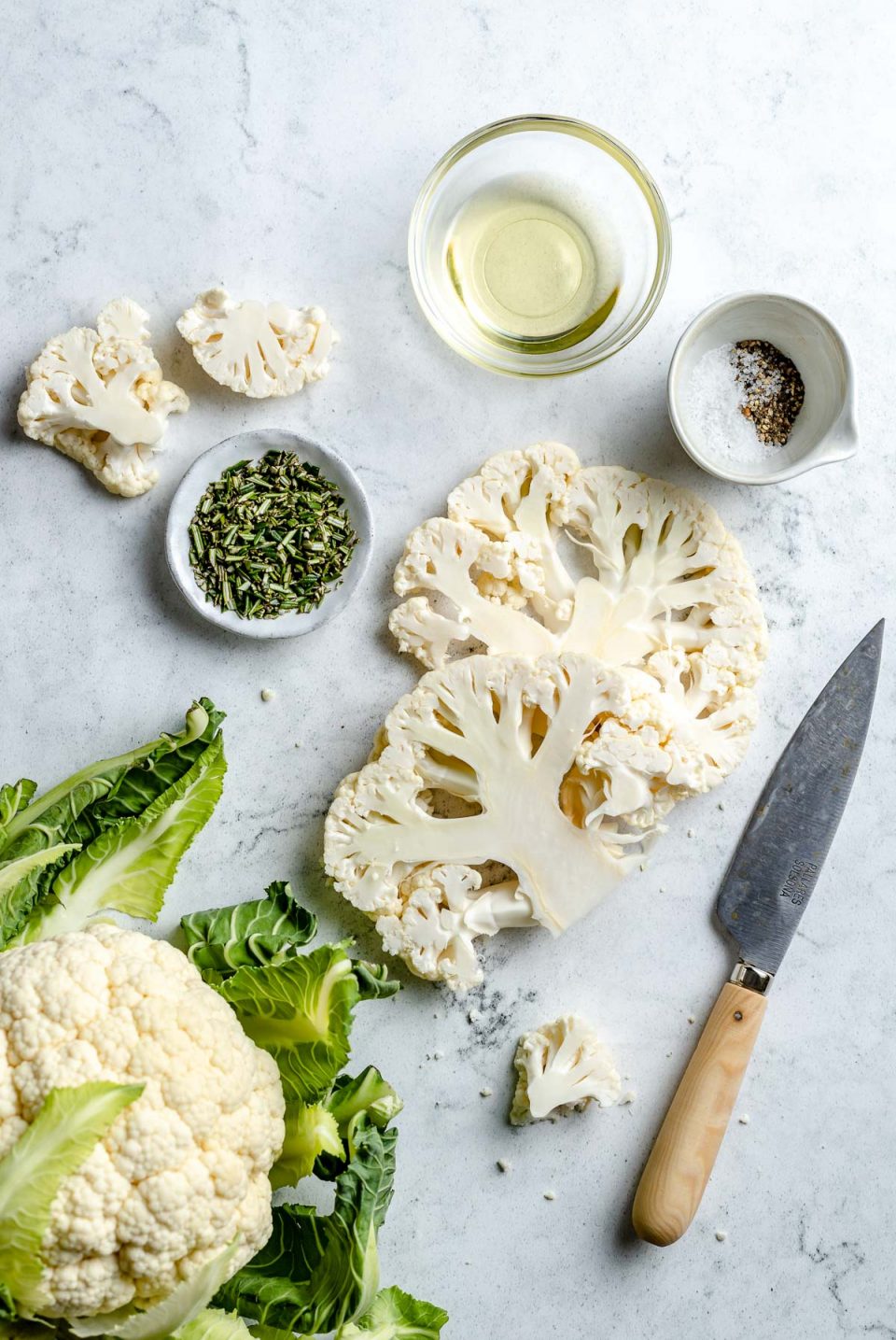 Grilled cauliflower ingredients arranged on a white & grey marble surface - a whole head of cauliflower, sliced cauliflower steaks & a few florets, avocado oil, kosher salt, ground black pepper, & chopped fresh herbs. A pairing knife rests on the surface next to the ingredients.