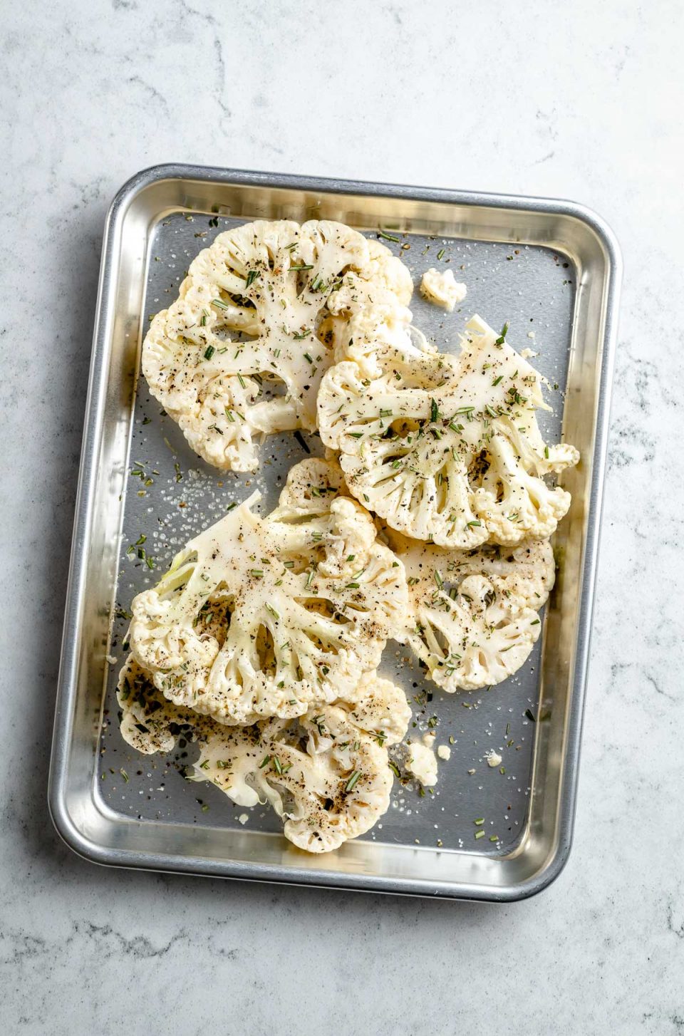 Multiple fresh & uncooked cauliflower steaks seasoned with oil, kosher salt, & ground black pepper lie on an aluminum baking sheet. The baking sheet sits on top of a white & gray marble surface.