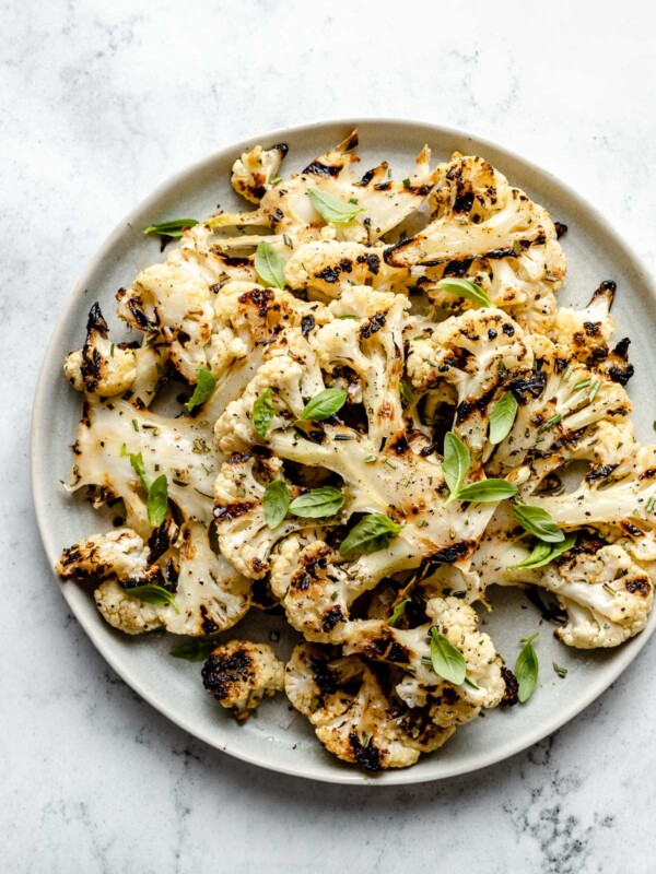 Grilled cauliflower with char marks arranged on a white ceramic plate. The cauliflower is garnished with ground black pepper, kosher salt, & fresh herbs. The plate sits on top of a white & gray marble surface.