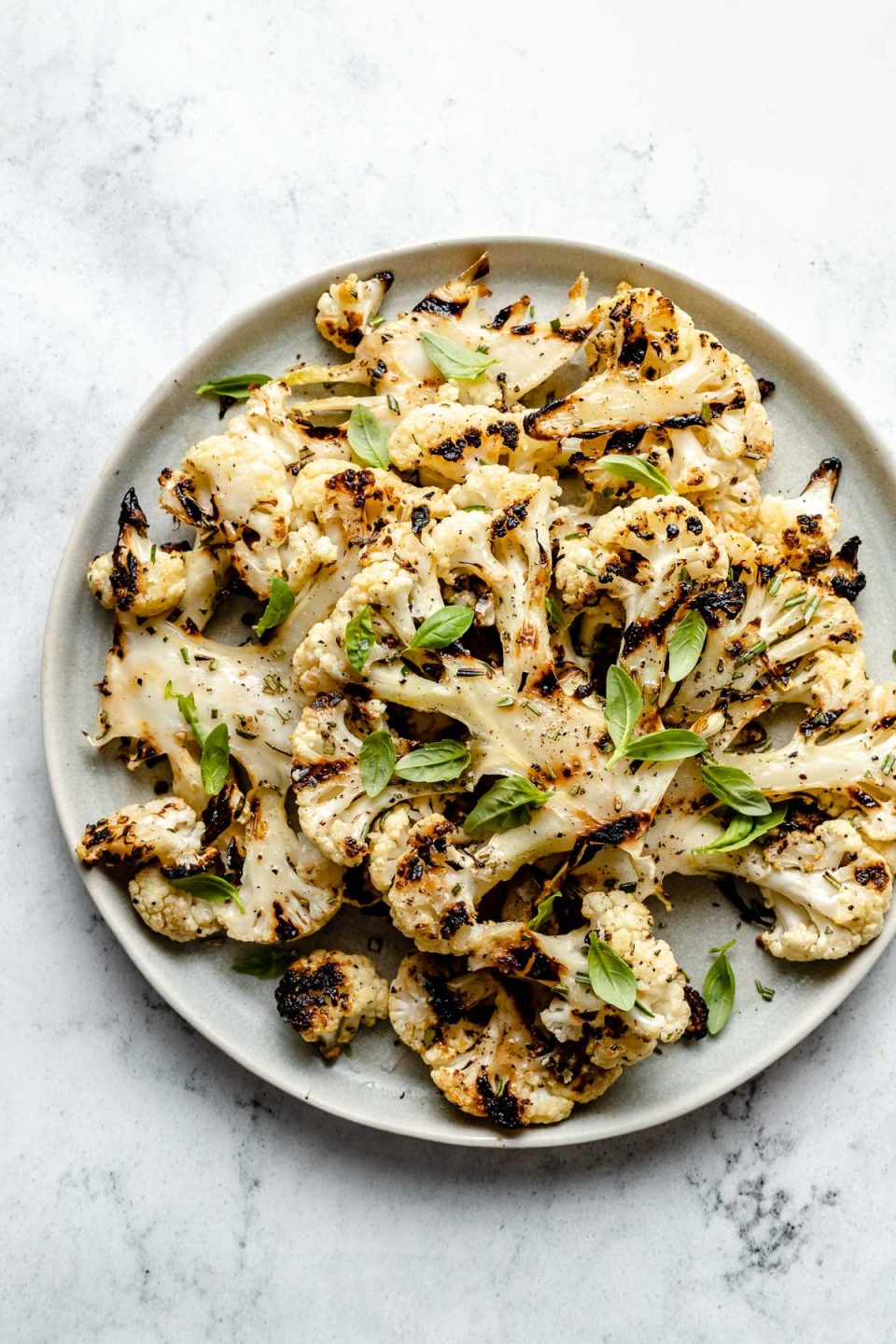Grilled cauliflower with char marks arranged on a white ceramic plate. The cauliflower is garnished with ground black pepper, kosher salt, & fresh herbs. The plate sits on top of a white & gray marble surface.