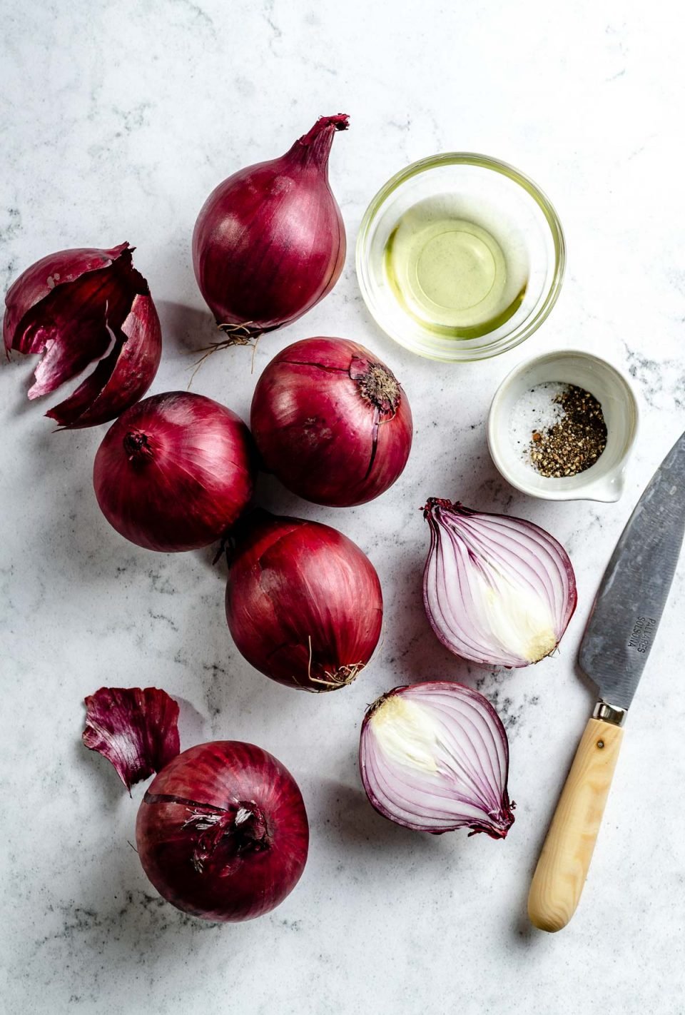 Grilled onion ingredients arranged on a white & grey marble surface - red onion, avocado oil, kosher salt, & ground black pepper. A pairing knife rests on the surface next to the ingredients.