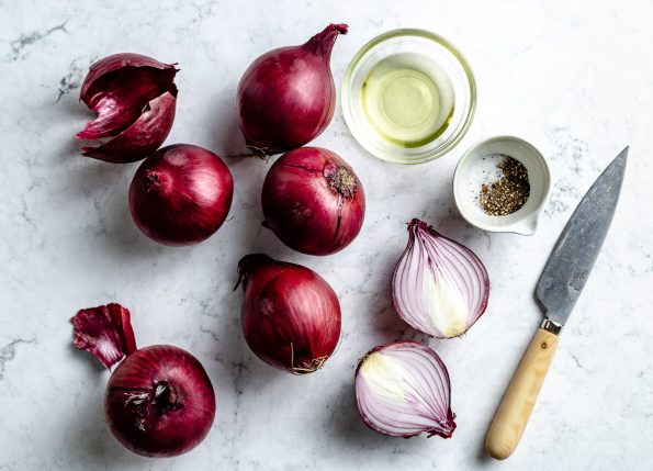 Grilled onion ingredients arranged on a white & grey marble surface - red onion, avocado oil, kosher salt, & ground black pepper. A pairing knife rests on the surface next to the ingredients.