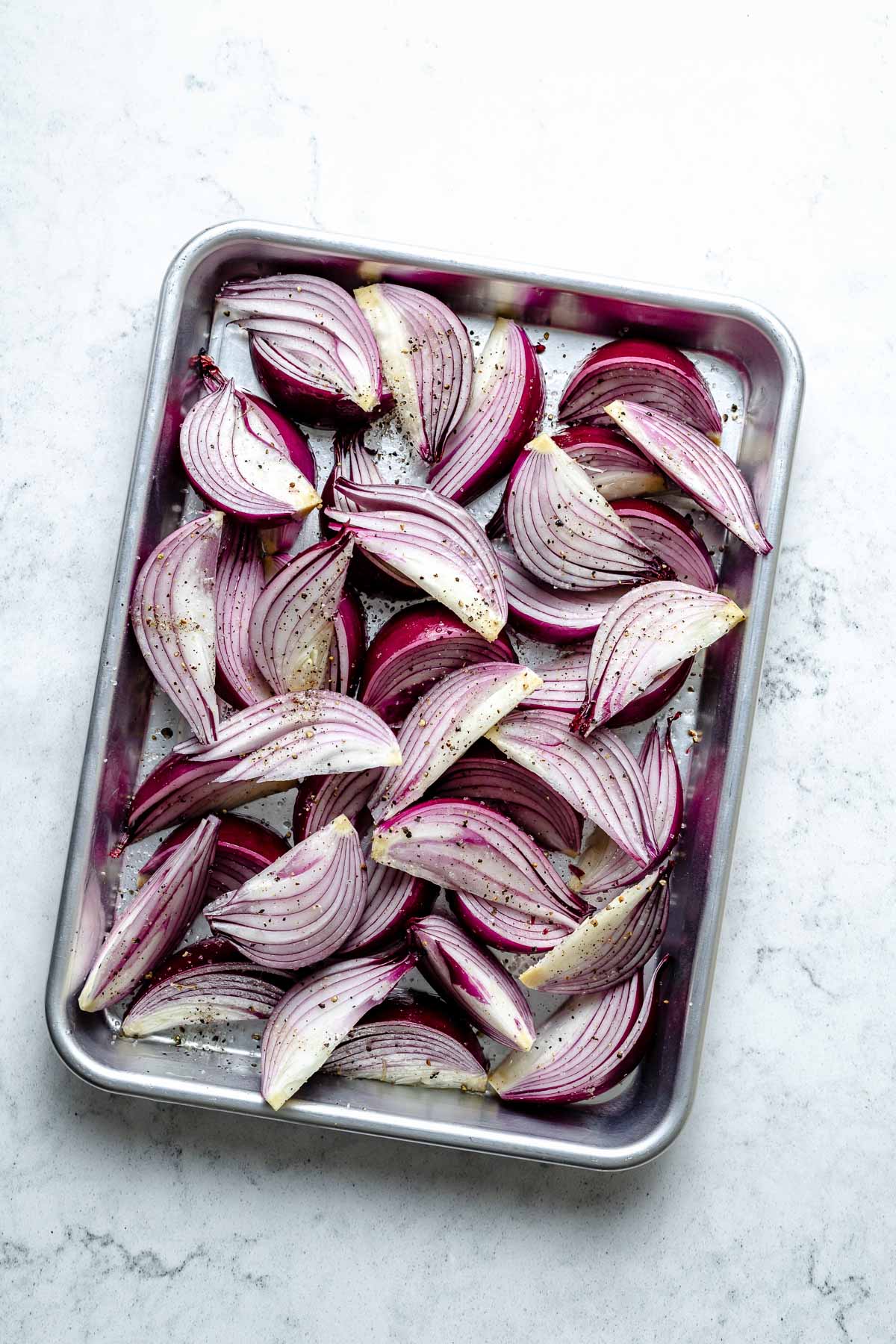 Multiple slices of red onion cut in wedges & seasoned with avocado oil, kosher salt, & ground black pepper arranged on an aluminum baking sheet. The baking sheet sits on top of a white & gray marble surface.